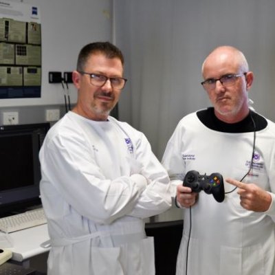 Two men in white lab coats in an office look at the camera, one holds a gaming controller. 